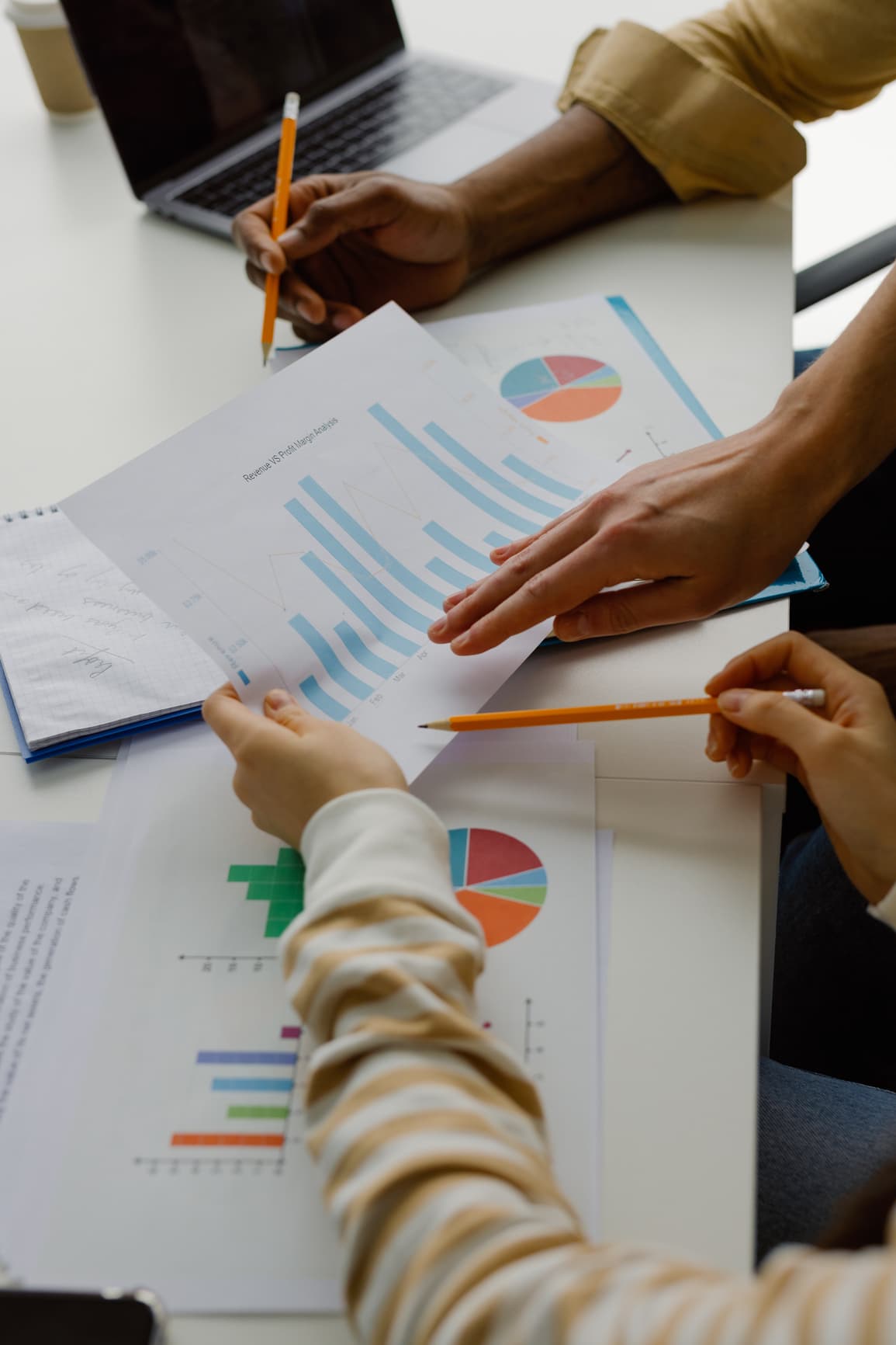 Two people pointing at and discussing printed charts and graphs on a desk with a laptop and coffee cup nearby.